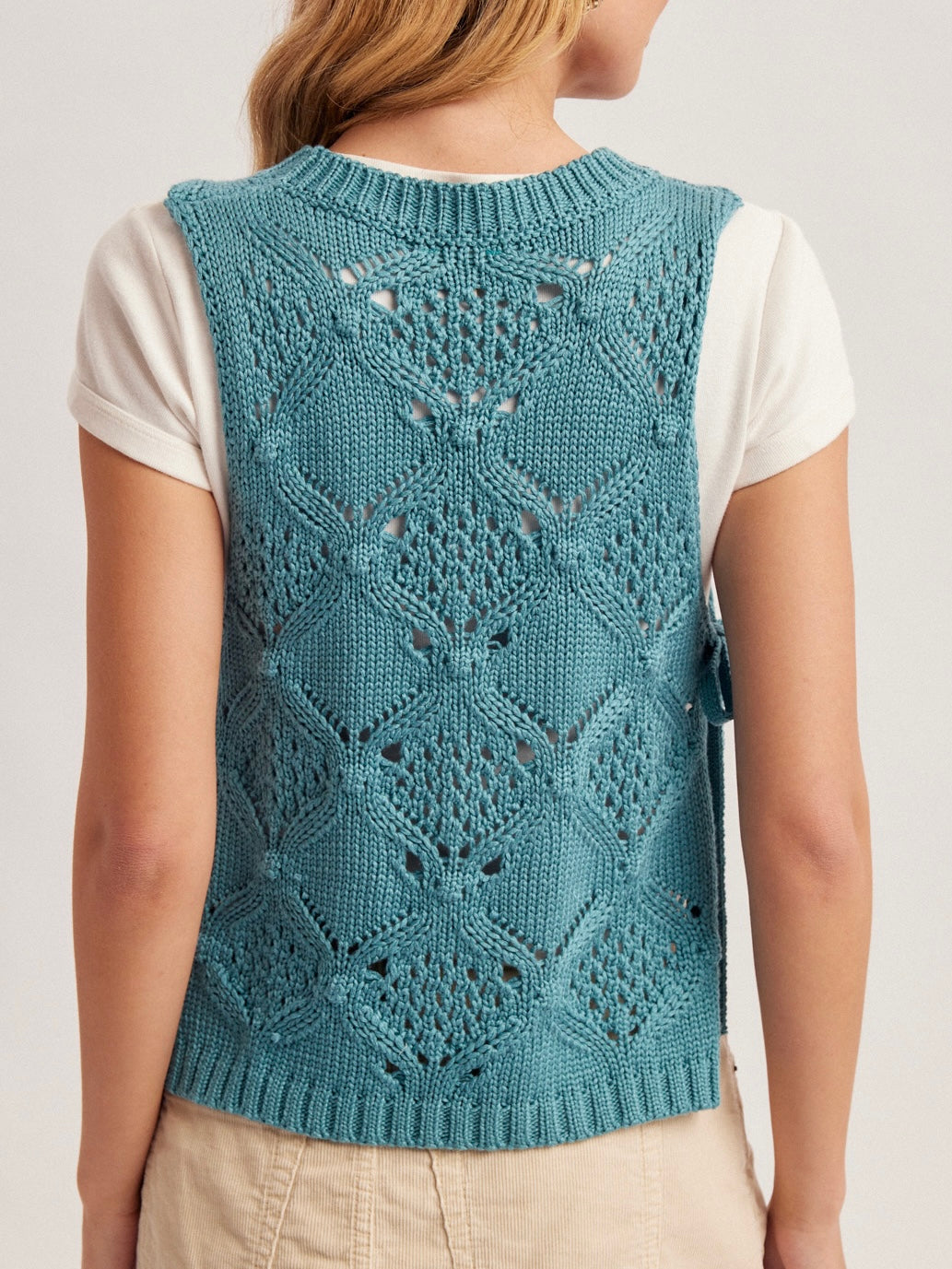 Teal knitted vest with a geometric pattern worn by a person on a light gray background