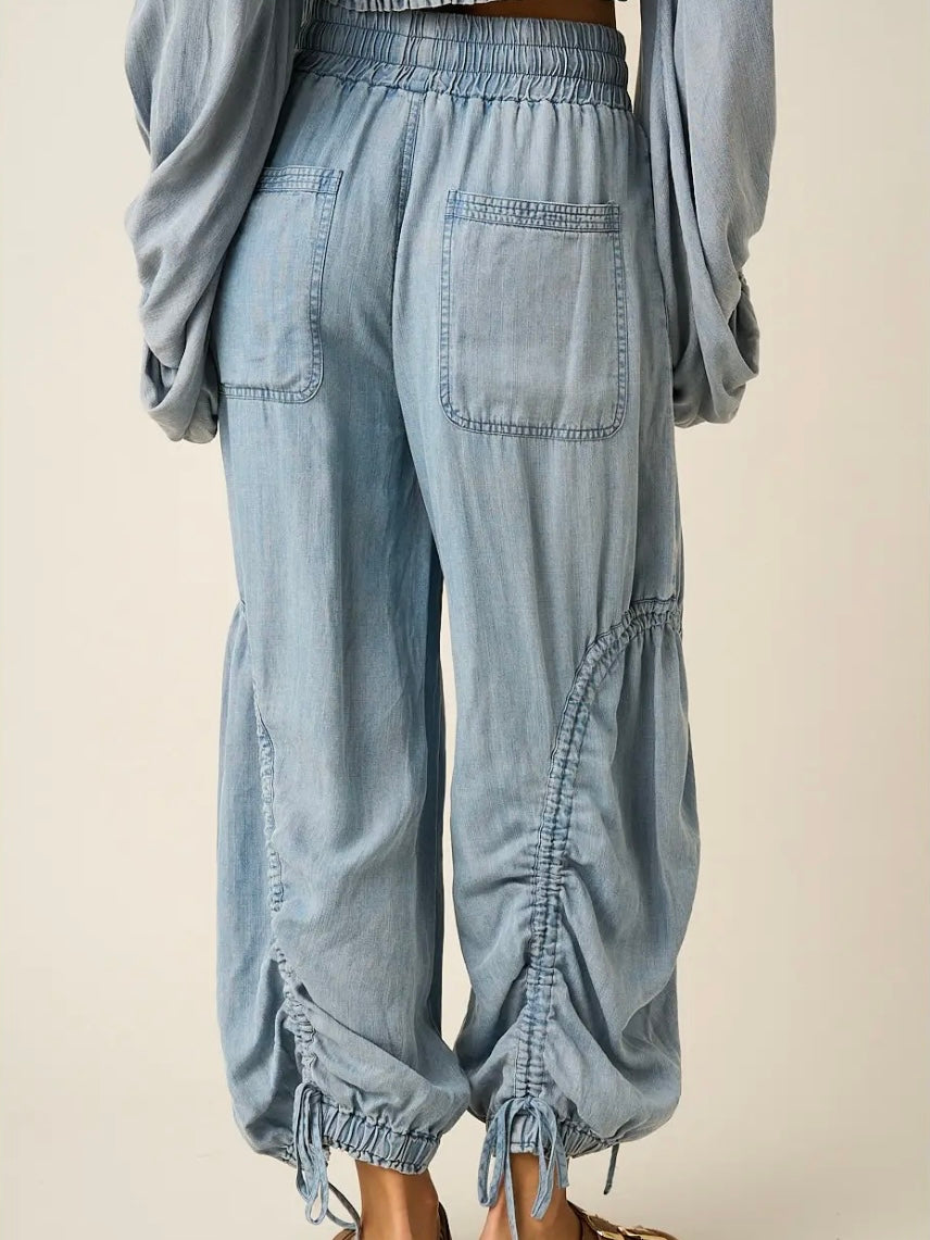 Light blue denim harem-style pants with an elastic waistband, large back patch pockets, gathered cuffed hems, and drawstring ties, shown on a model from the back against a plain beige background.