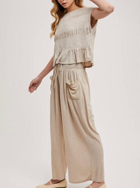 A woman modeling a two-piece beige knit set consisting of a sleeveless smocked ruffle crop top and matching wide-leg pants with side pockets, set against a plain light gray studio background.