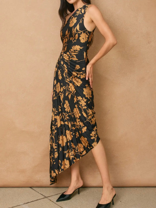 A woman models a sleeveless, high-neck, midi-length dress with a black base and a warm brown floral pattern, paired with black pointed-toe mule heels against a textured beige wall.