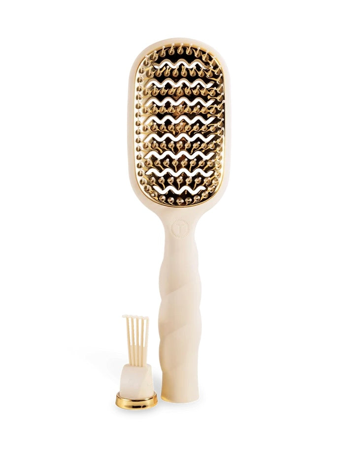 A cream-colored hairbrush with gold metallic bristles, shown alongside its small gold and cream cleaning brush accessory on a solid white background.