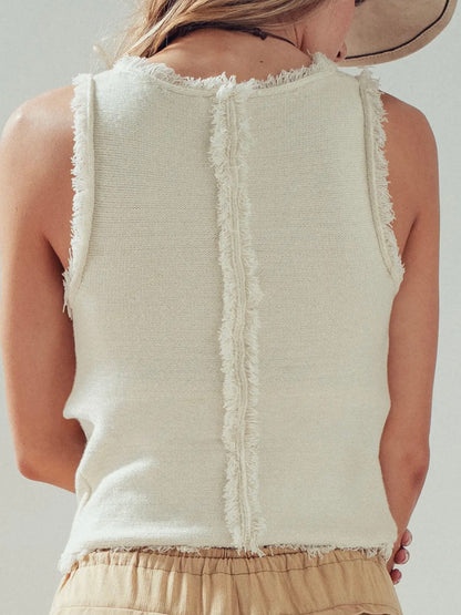Person wearing a beige sleeveless top with fringe details on a plain background