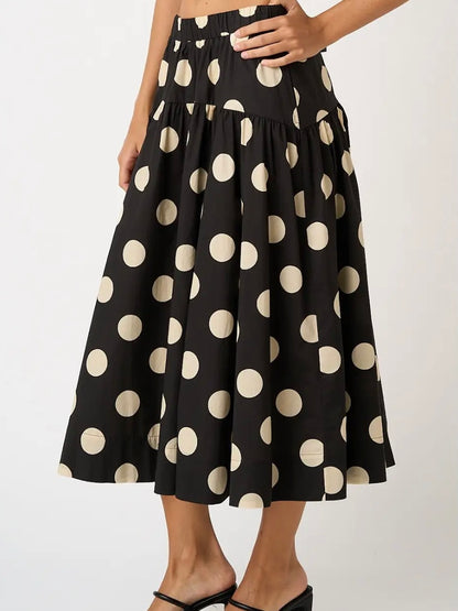 A black midi skirt with a large beige polka dot pattern, shown on a model wearing black strappy heels against a light gray studio background.