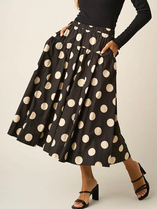 A black midi skirt with large beige polka dots, styled with a black ribbed long-sleeve top and black strappy heels against a light beige studio background.