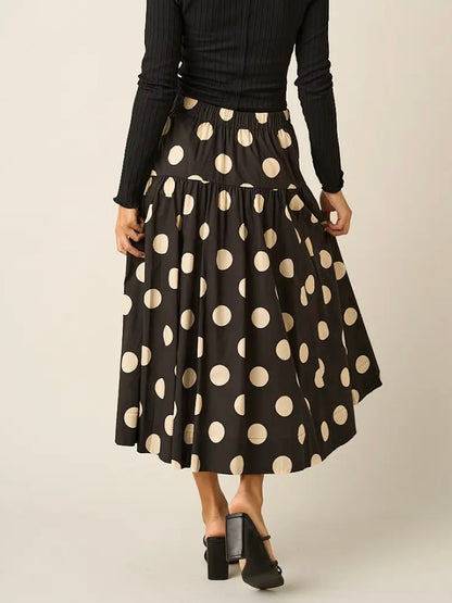 A model wearing a black midi skirt with large beige polka dots, paired with a black ribbed long-sleeve top and black block-heel mules, shown from the back against a light beige studio background.