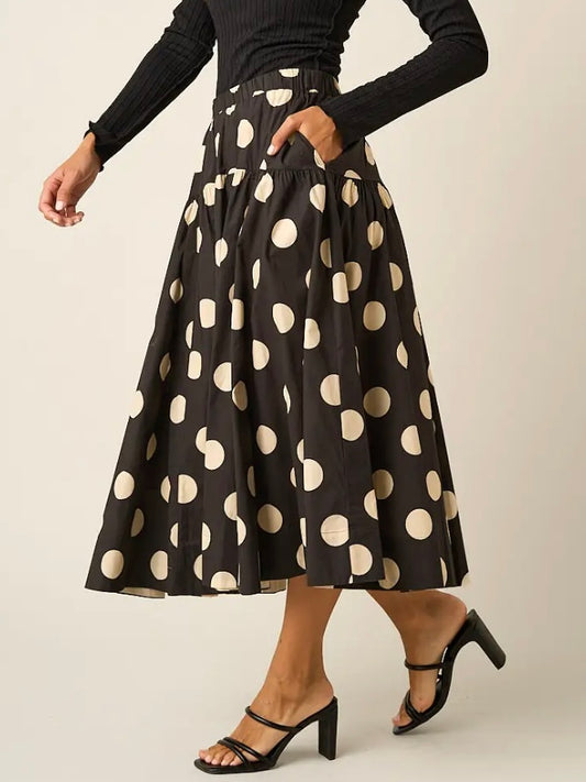 A model wearing a black midi skirt with large beige polka dots, paired with a black long-sleeve top and black heeled sandals, against a light beige studio background.