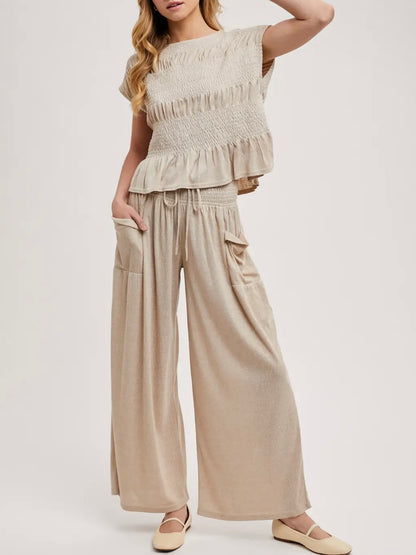 A woman modeling a light beige two-piece outfit consisting of a short-sleeve smocked peplum top and matching wide-leg pants with side pockets, paired with beige flats, against a plain light gray studio background.