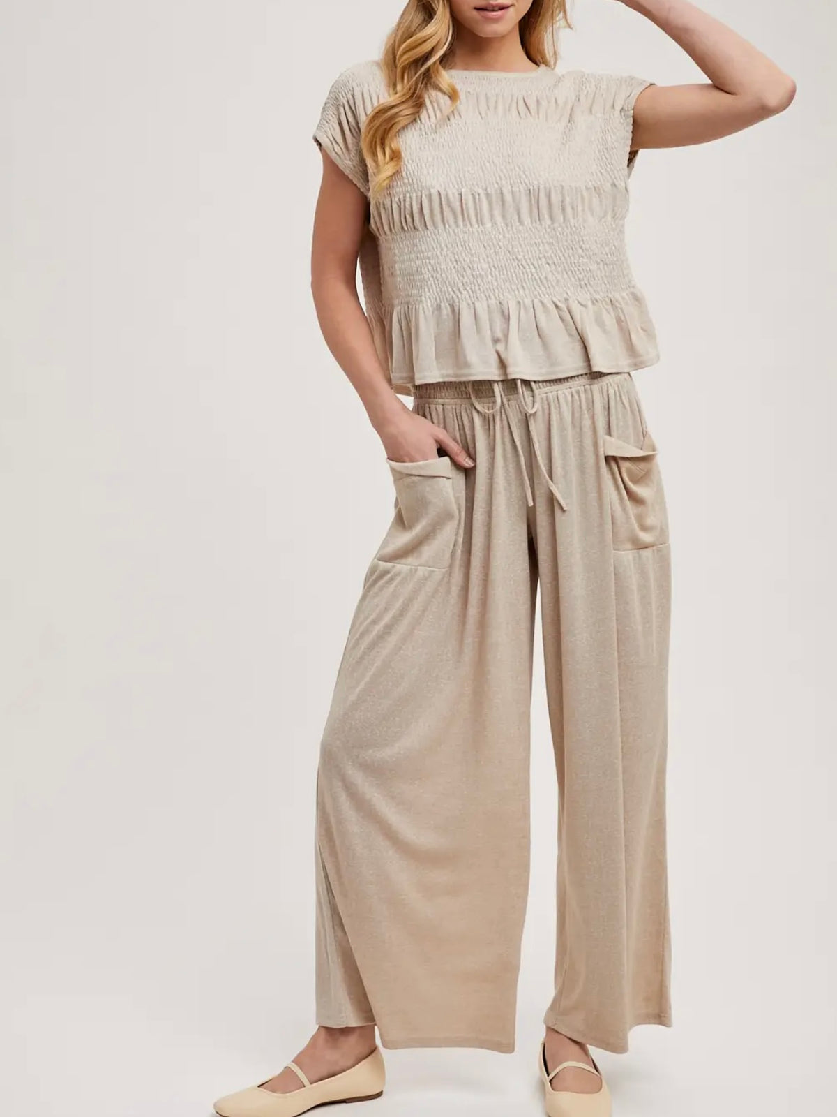 A woman modeling a beige two-piece lounge set consisting of a short-sleeve smocked ruffle top and matching wide-leg drawstring pants with side pockets, paired with beige flats against a light gray studio background.