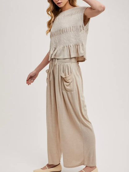 A woman modeling a two-piece beige knit set consisting of a sleeveless smocked ruffle crop top and matching wide-leg pants with side pockets, set against a plain light gray studio background.