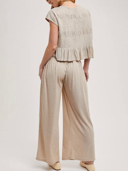 A woman shown from the back wearing a light beige two-piece set consisting of a short-sleeve cropped top with horizontal ruching and ruffle trim, paired with matching wide-leg pants, against a plain light background.
