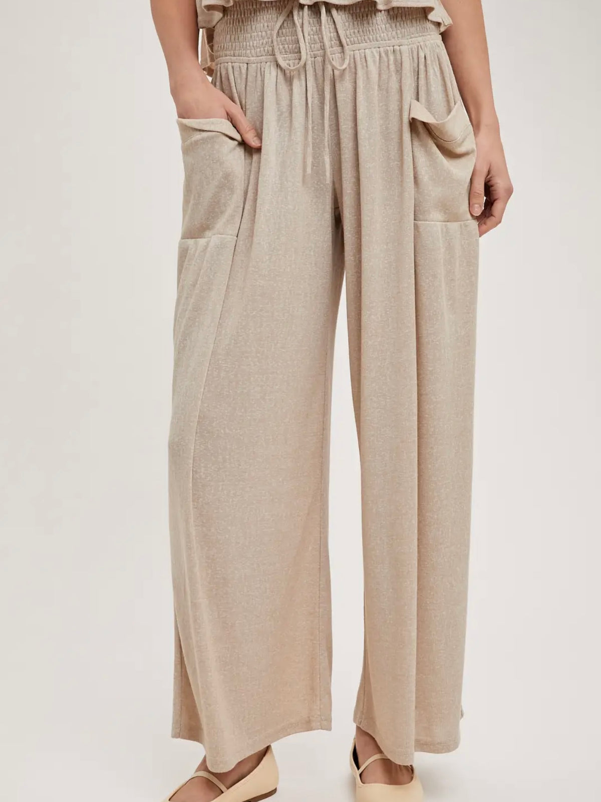 A person wearing loose, wide-leg beige pants with a smocked waistband, drawstring, and large front patch pockets, shown against a plain light background.