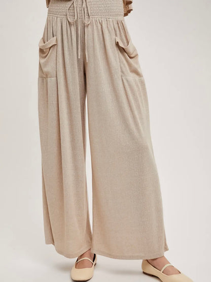 A person wearing loose, wide-leg beige pants with a smocked waistband, drawstring, and large front patch pockets, paired with light beige flats, against a plain light background.