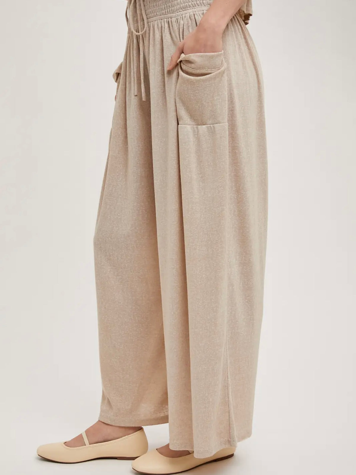 A person wearing loose, wide-leg beige pants with a drawstring waist, side pockets, and a smocked waistband, paired with beige flats against a plain light background.