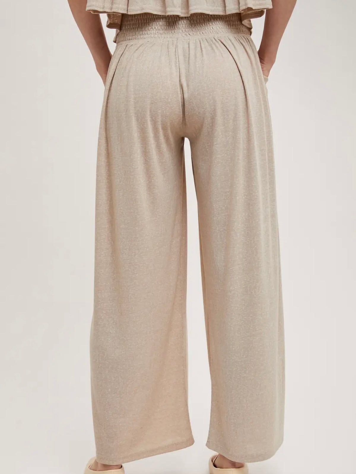 A person shown from the back wearing loose, wide-leg pleated pants in a light warm beige or oatmeal tone, against a plain light gray studio background.