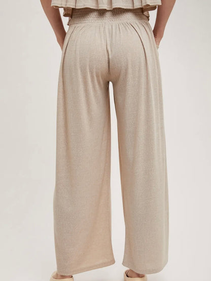 A person shown from the back wearing loose, wide-leg pleated pants in a light warm beige or oatmeal tone, against a plain light gray studio background.