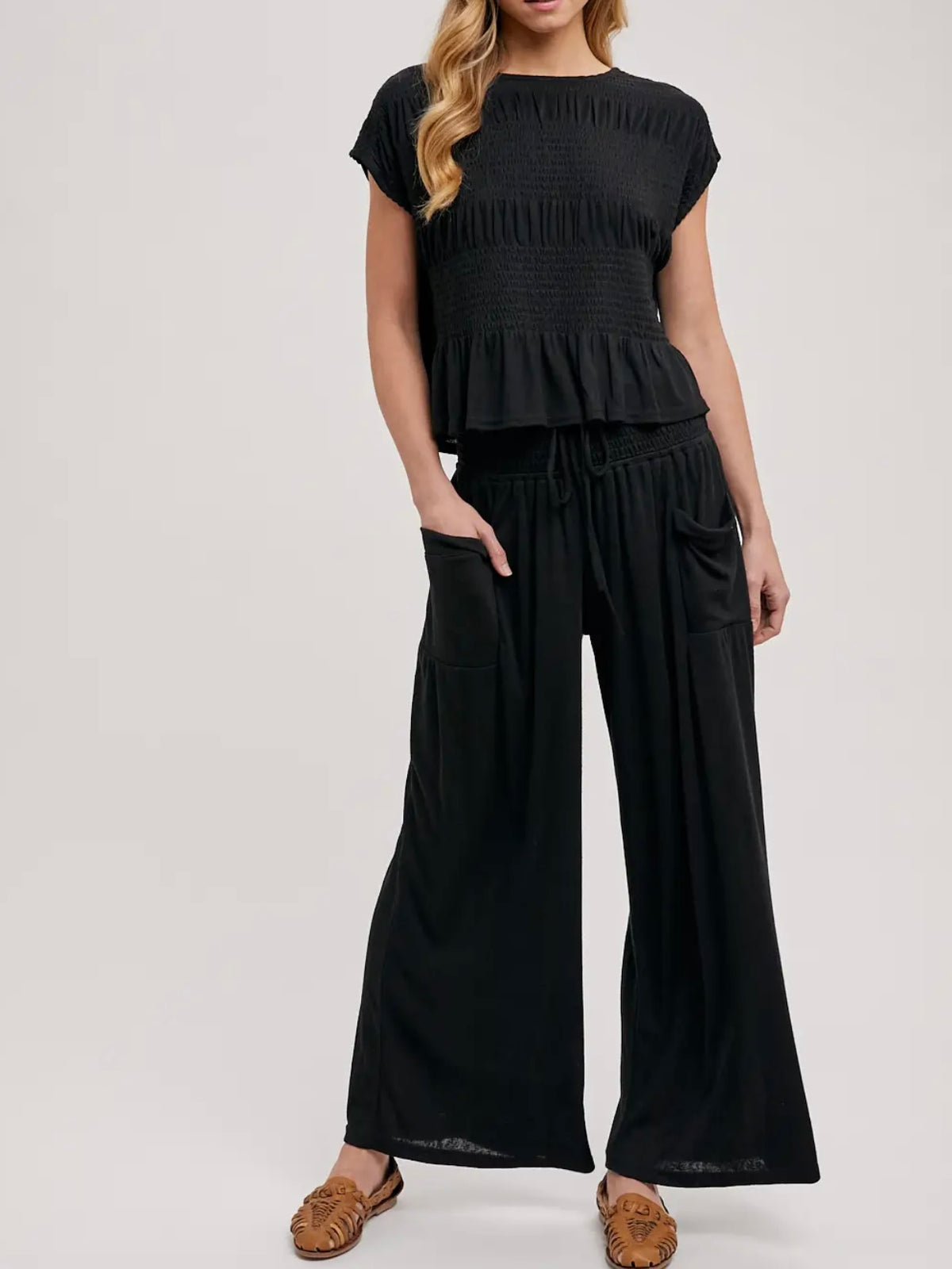 A woman modeling a black two-piece outfit consisting of a short-sleeve, smocked-peplum top and matching wide-leg pants, paired with brown flat shoes against a light gray studio background.