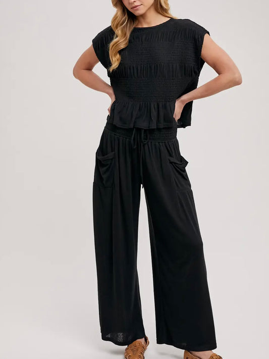 A woman modeling a black two-piece outfit consisting of a short-sleeve, smocked, and ruffled crop top paired with high-waisted, wide-leg pants featuring side pockets, set against a plain light gray studio background.