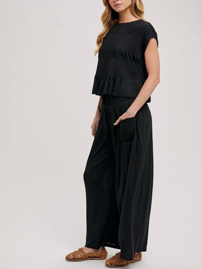 A woman modeling a black two-piece outfit consisting of a short-sleeve ruffled crop top and matching wide-leg pants, paired with brown flat sandals, against a plain light gray studio background.