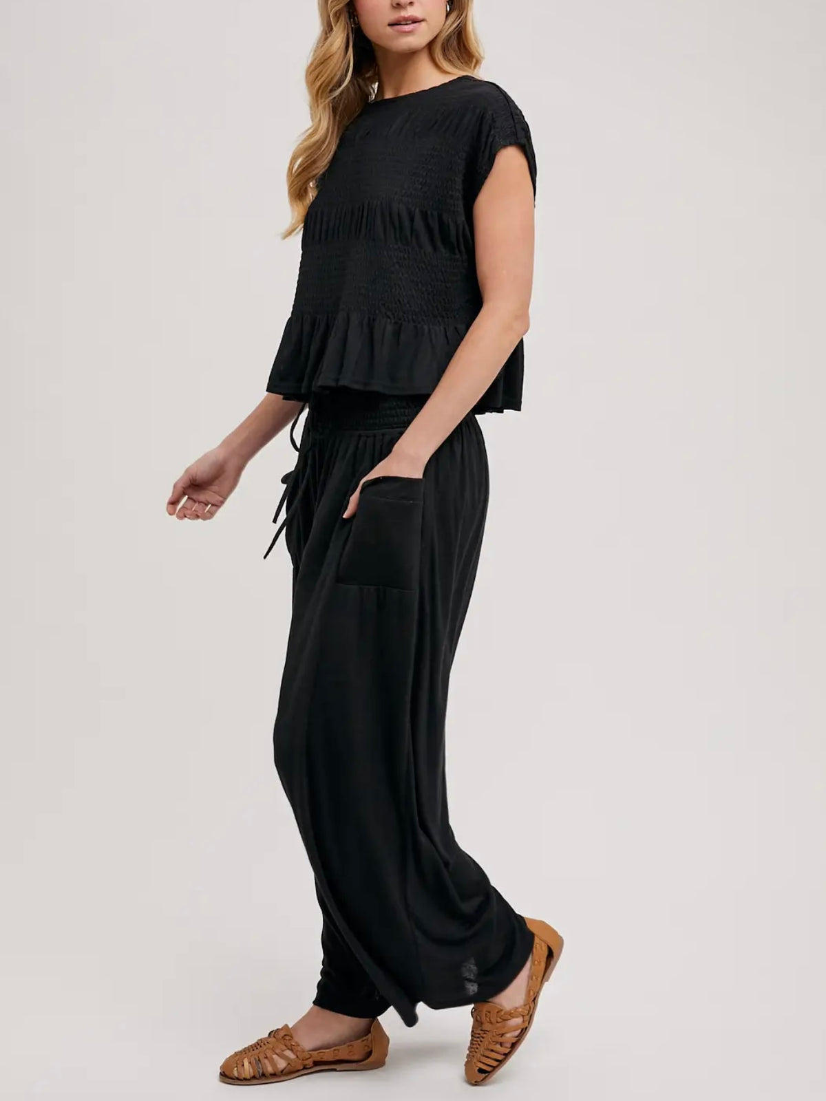 A woman modeling a black two-piece outfit consisting of a short-sleeve ruffled crop top and matching loose black drawstring pants, paired with brown leather flat sandals, against a plain light gray studio background.