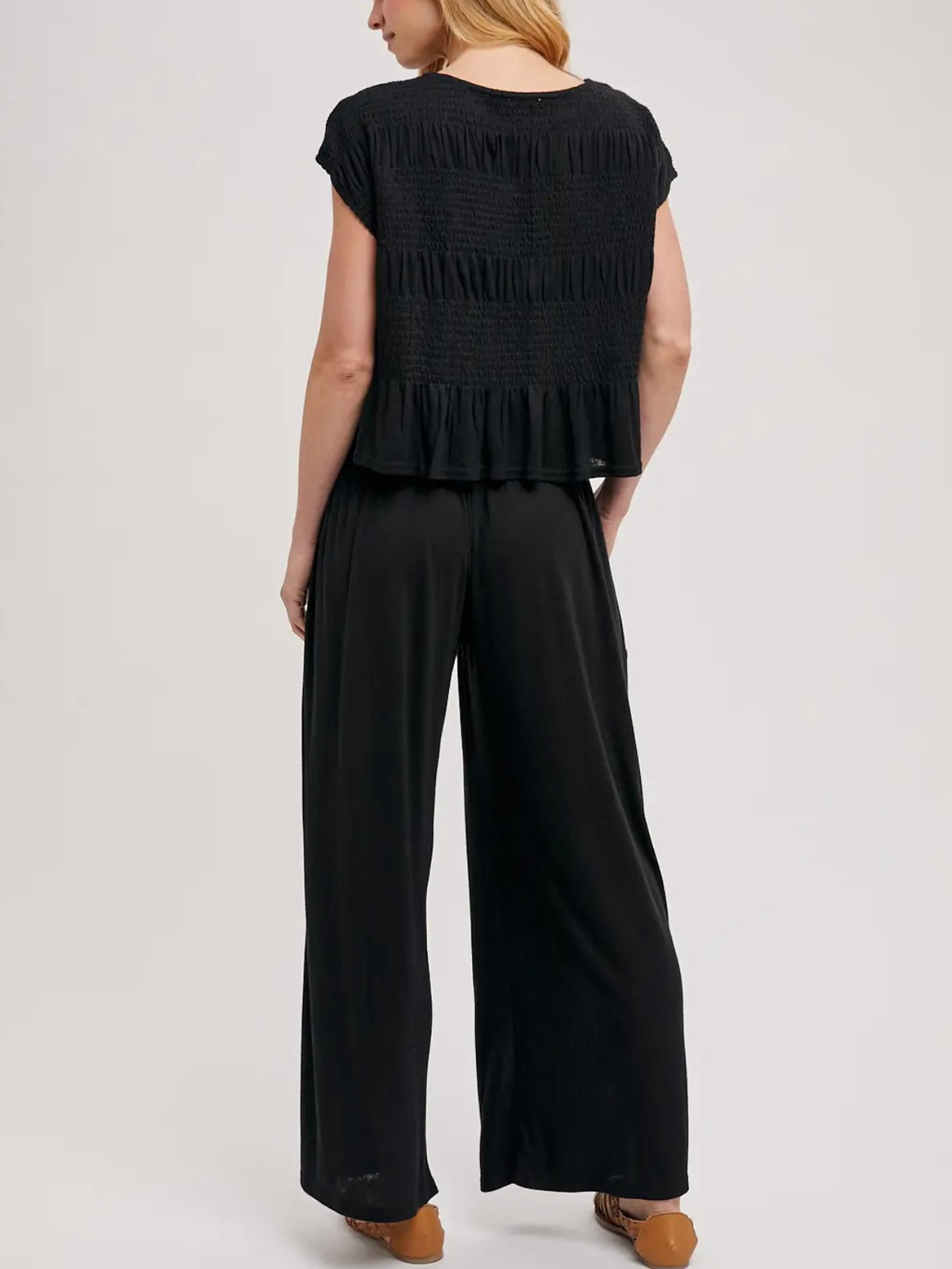 A woman shown from the back wearing a black smocked short-sleeve top with a ruffled hem and black wide-leg pants, photographed against a light gray studio background.