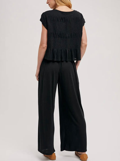 A woman shown from the back wearing a black smocked short-sleeve top with a ruffled hem and black wide-leg pants, photographed against a light gray studio background.