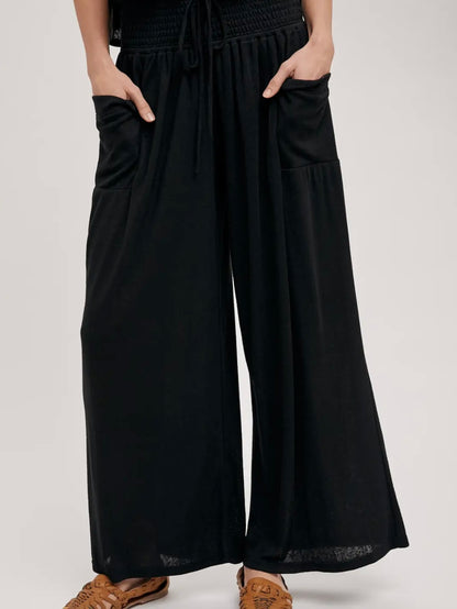 Black wide-leg pants with a smocked elastic waist, drawstring, and large front patch pockets, shown on a model wearing brown sandals against a light gray studio background.