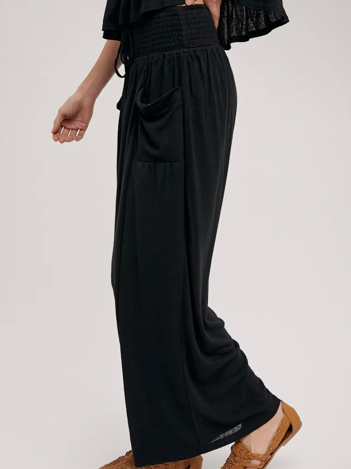 A person wearing loose black wide-leg pants with a smocked waistband, side pockets, and a drawstring, paired with brown flat shoes against a light gray studio background.