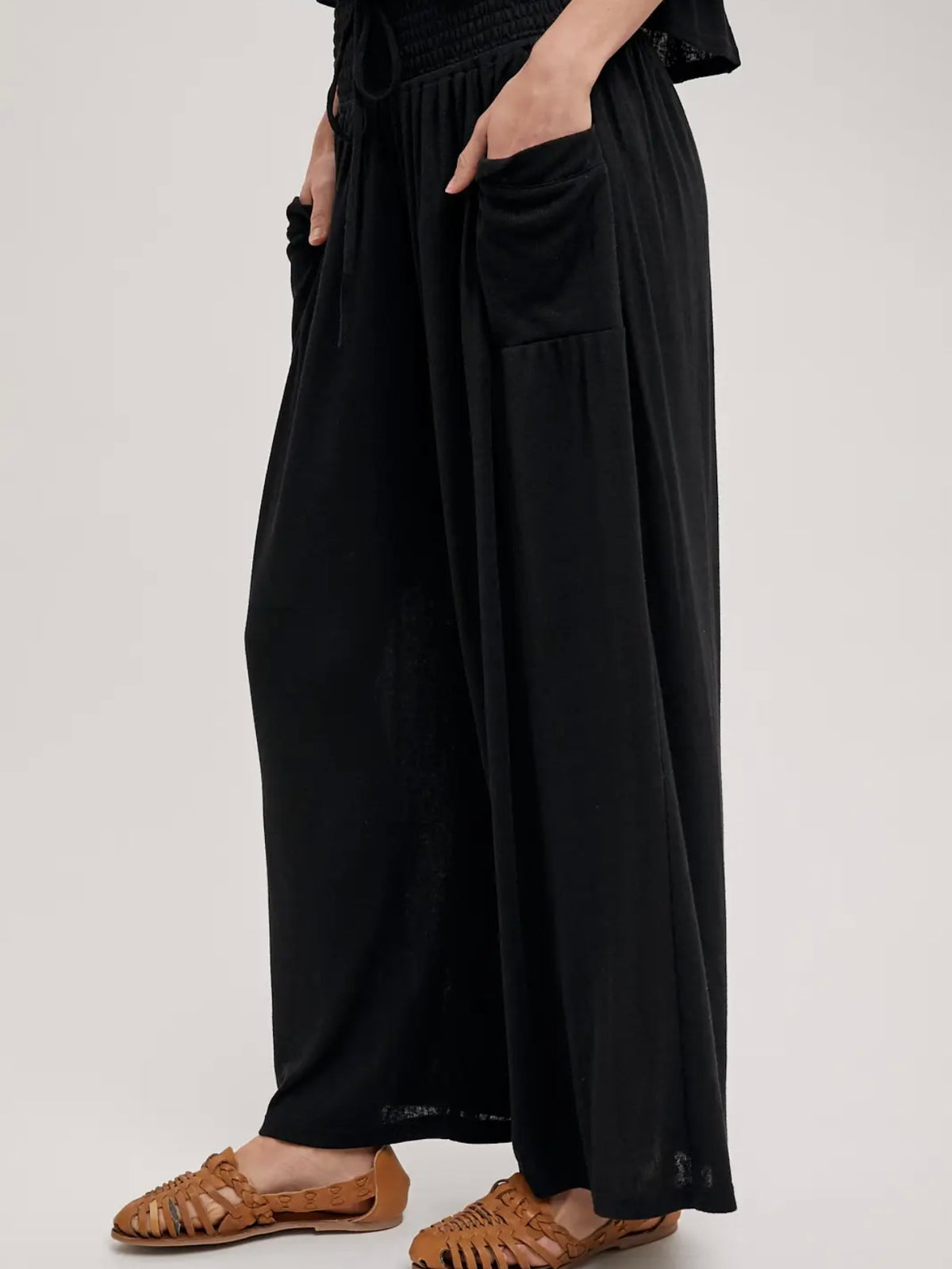 A person wearing loose black wide-leg pants with side pockets and brown woven sandals, shown from the waist down against a plain light gray studio background.