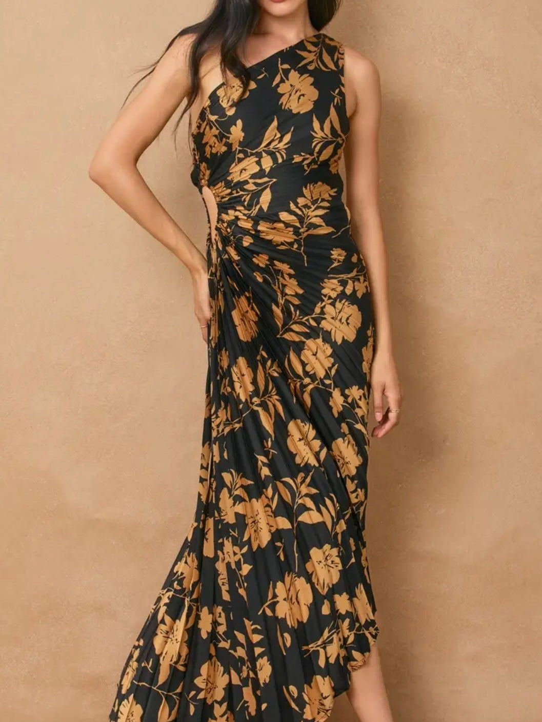 A woman wearing a sleeveless one-shoulder maxi dress with a black base and a large tan floral print, posed against a textured beige wall.