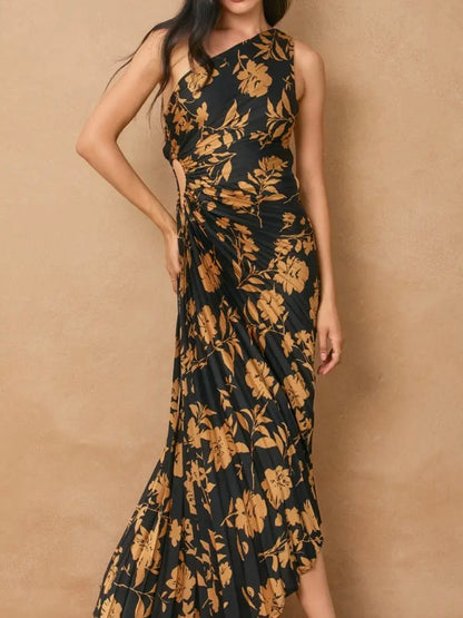 A woman wearing a sleeveless one-shoulder maxi dress with a black base and a large tan floral print, posed against a textured beige wall.