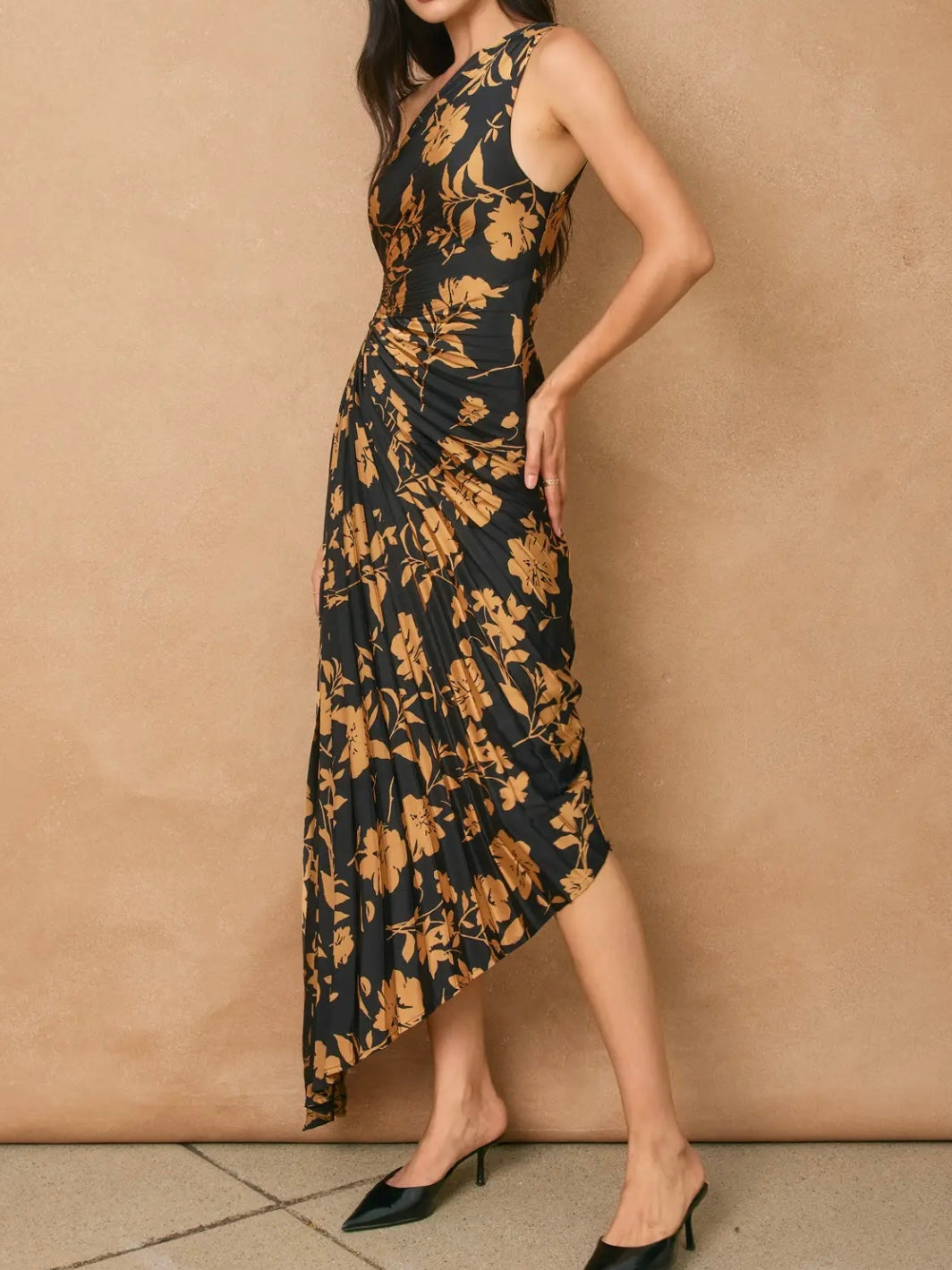 A woman models a sleeveless, high-neck, midi-length dress with a black base and a warm brown floral pattern, paired with black pointed-toe mule heels against a textured beige wall.