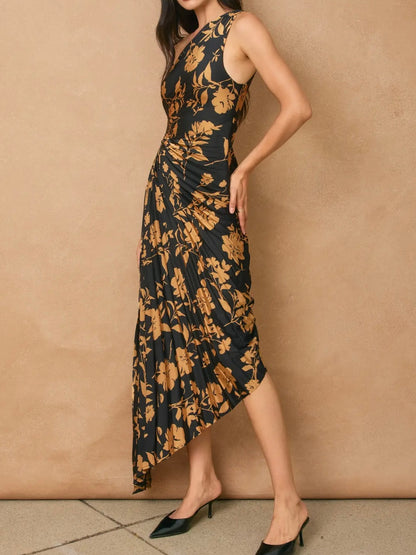 A woman models a sleeveless, high-neck, midi-length dress with a black base and a warm brown floral pattern, paired with black pointed-toe mule heels against a textured beige wall.