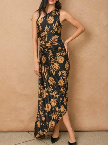 A woman models a sleeveless, one-shoulder maxi dress with a black base and a prominent mustard yellow floral pattern, posed against a textured beige wall.