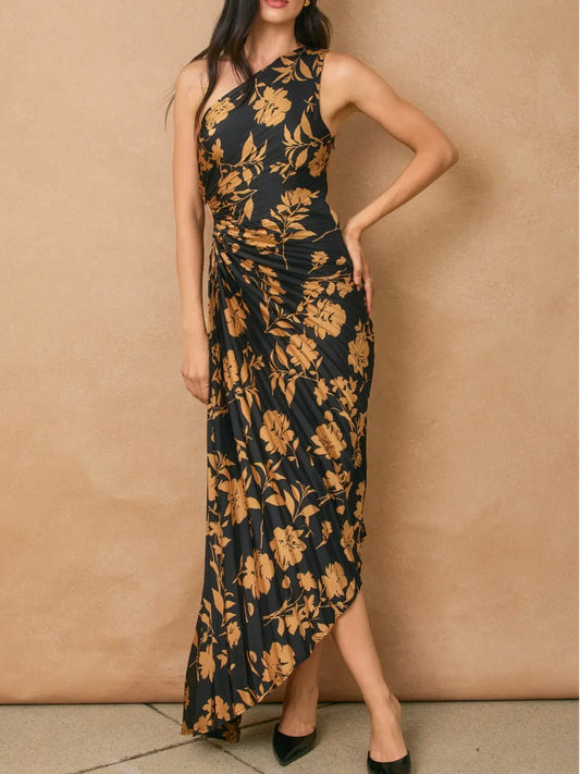 A woman models a sleeveless, one-shoulder maxi dress with a black base and a prominent mustard yellow floral pattern, posed against a textured beige wall.