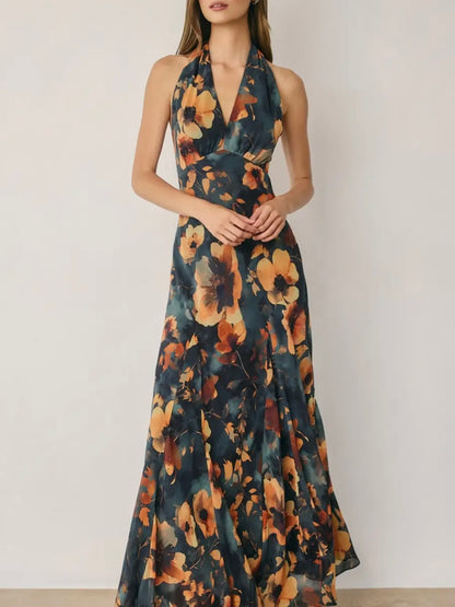 A woman wearing a sleeveless halter-neck maxi dress with a dark teal and orange floral watercolor print, standing against a plain off-white wall.