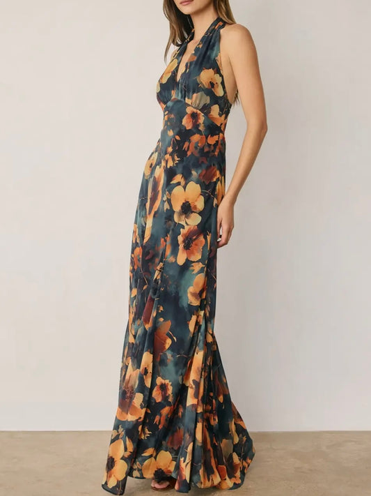 A woman modeling a sleeveless halter-neck maxi dress with a dark teal background and a large orange and yellow floral print, set against a plain light beige wall.