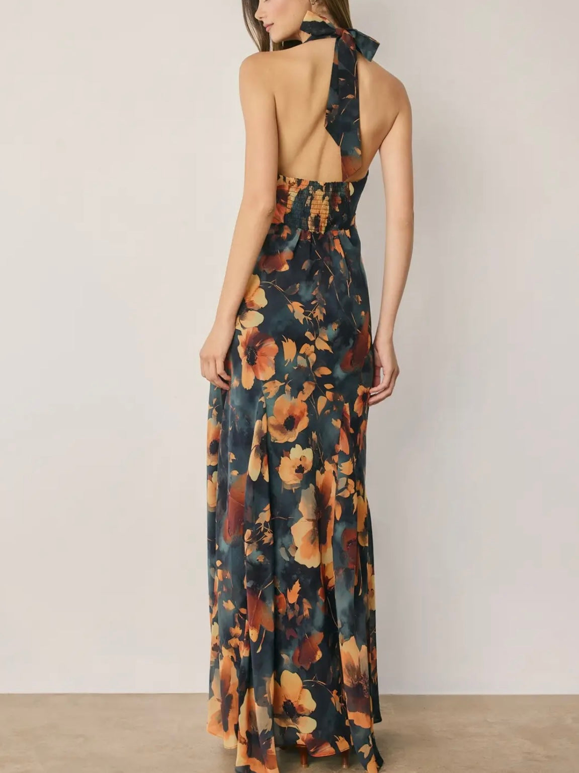 A woman modeling a halter-neck maxi dress from the back, featuring a dark navy blue base with a large orange and yellow floral print, set against a plain light gray wall.