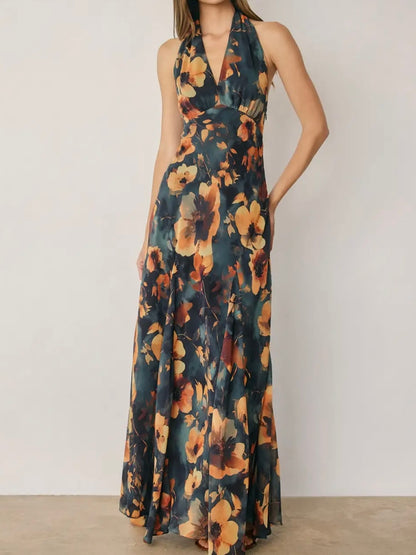 A woman modeling a sleeveless halter-neck maxi dress with a dark teal floral print featuring large orange and yellow flowers, set against a plain off-white wall.