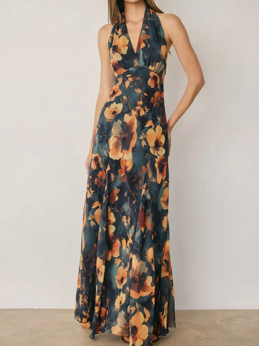 A woman modeling a sleeveless halter-neck maxi dress with a dark teal floral print featuring large orange and yellow flowers, set against a plain off-white wall.