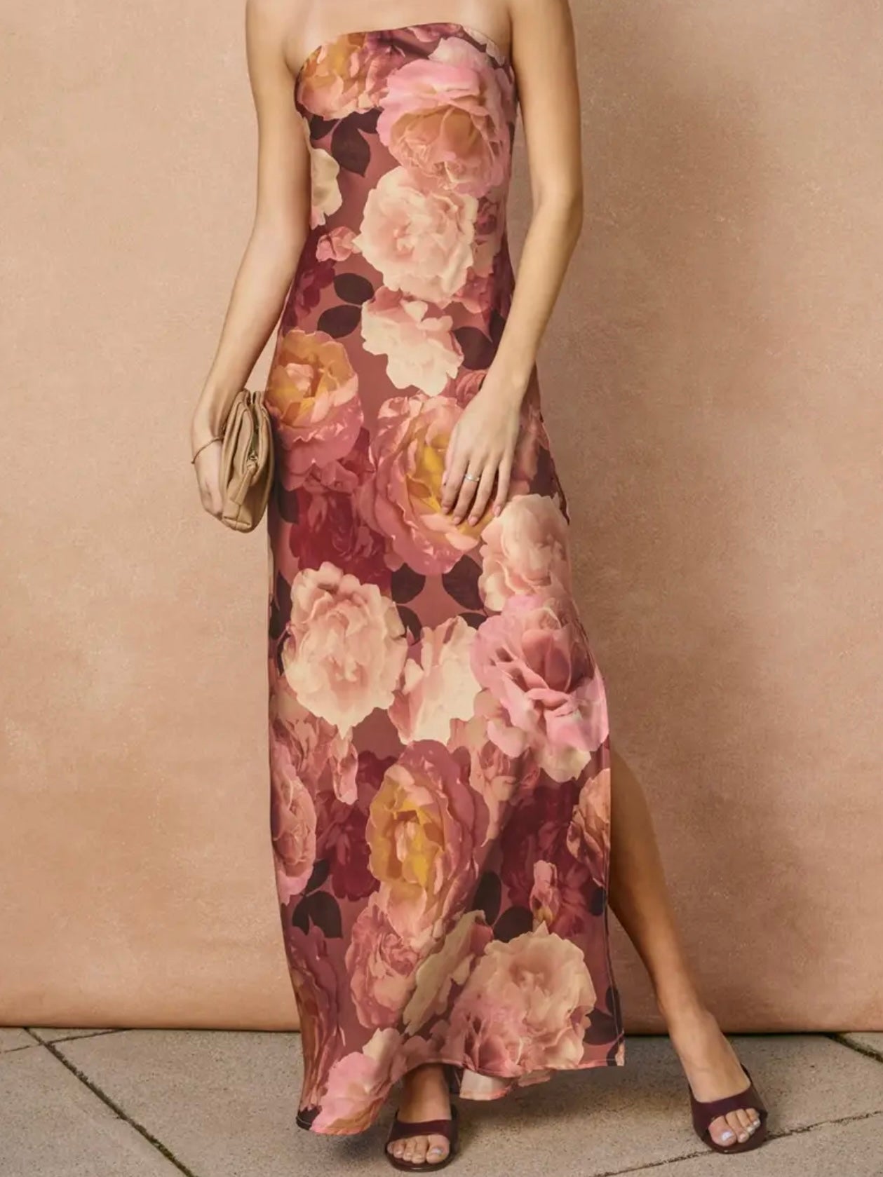 A woman models a strapless maxi dress with a large floral print in shades of pink, mauve, and yellow, paired with dark purple heeled sandals and a small beige clutch, set against a textured beige wall.