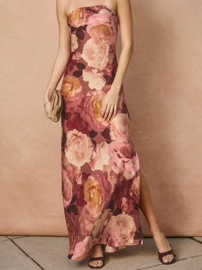 A woman models a strapless maxi dress with a large floral print in shades of pink, mauve, and yellow, paired with dark purple heeled sandals and a small beige clutch, set against a textured beige wall.