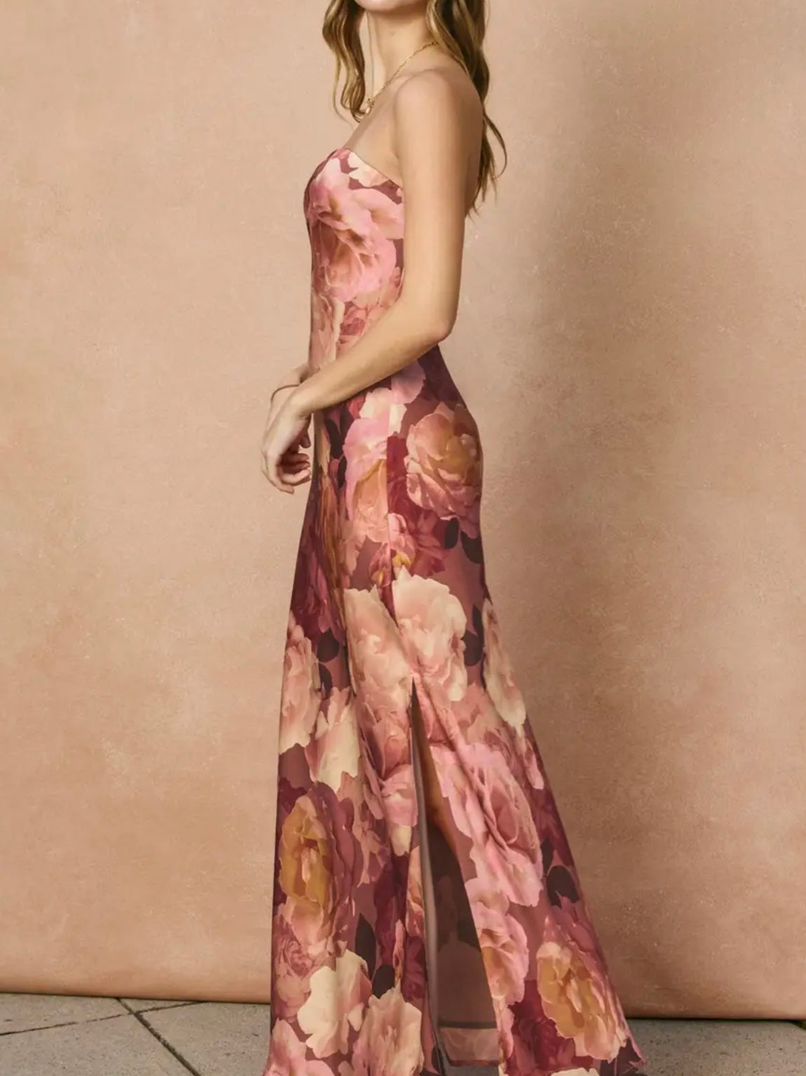 A woman wearing a sleeveless, floor-length floral gown with a thigh-high slit, posed in profile against a warm beige textured backdrop.