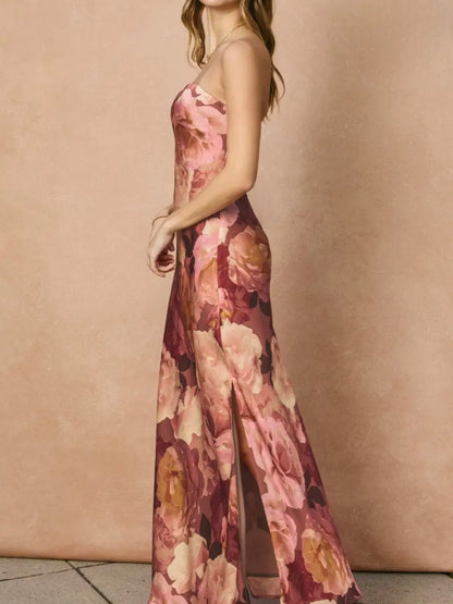 A woman wearing a sleeveless, floor-length floral gown with a thigh-high slit, posed in profile against a warm beige textured backdrop.