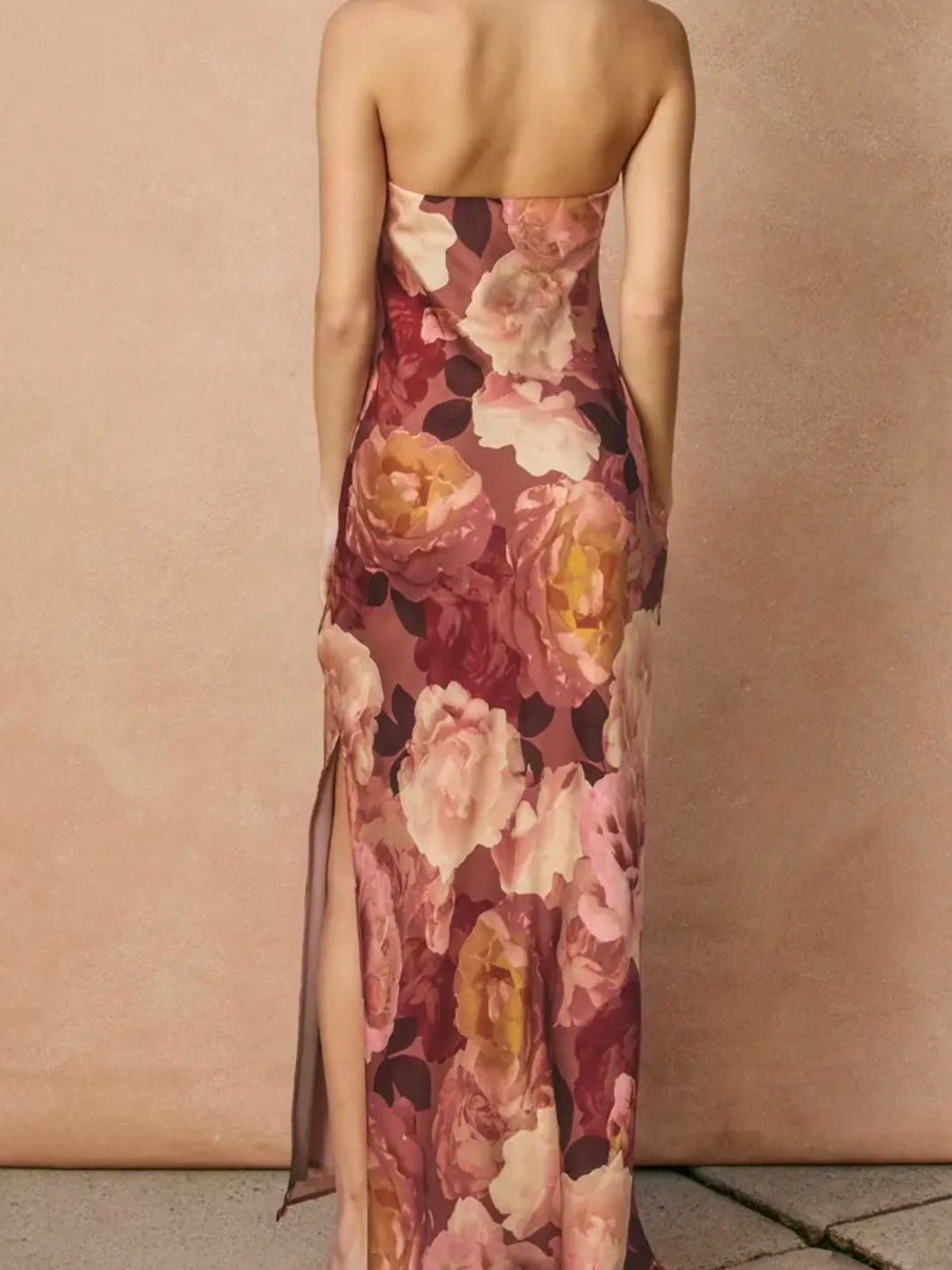 A strapless floral maxi dress with a side slit, shown from the back on a model against a textured beige wall.