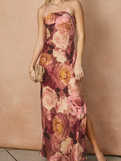 A woman models a strapless maxi dress with a large floral print in dusty pink, mauve, and yellow tones, holding a small beige clutch against a textured beige wall.