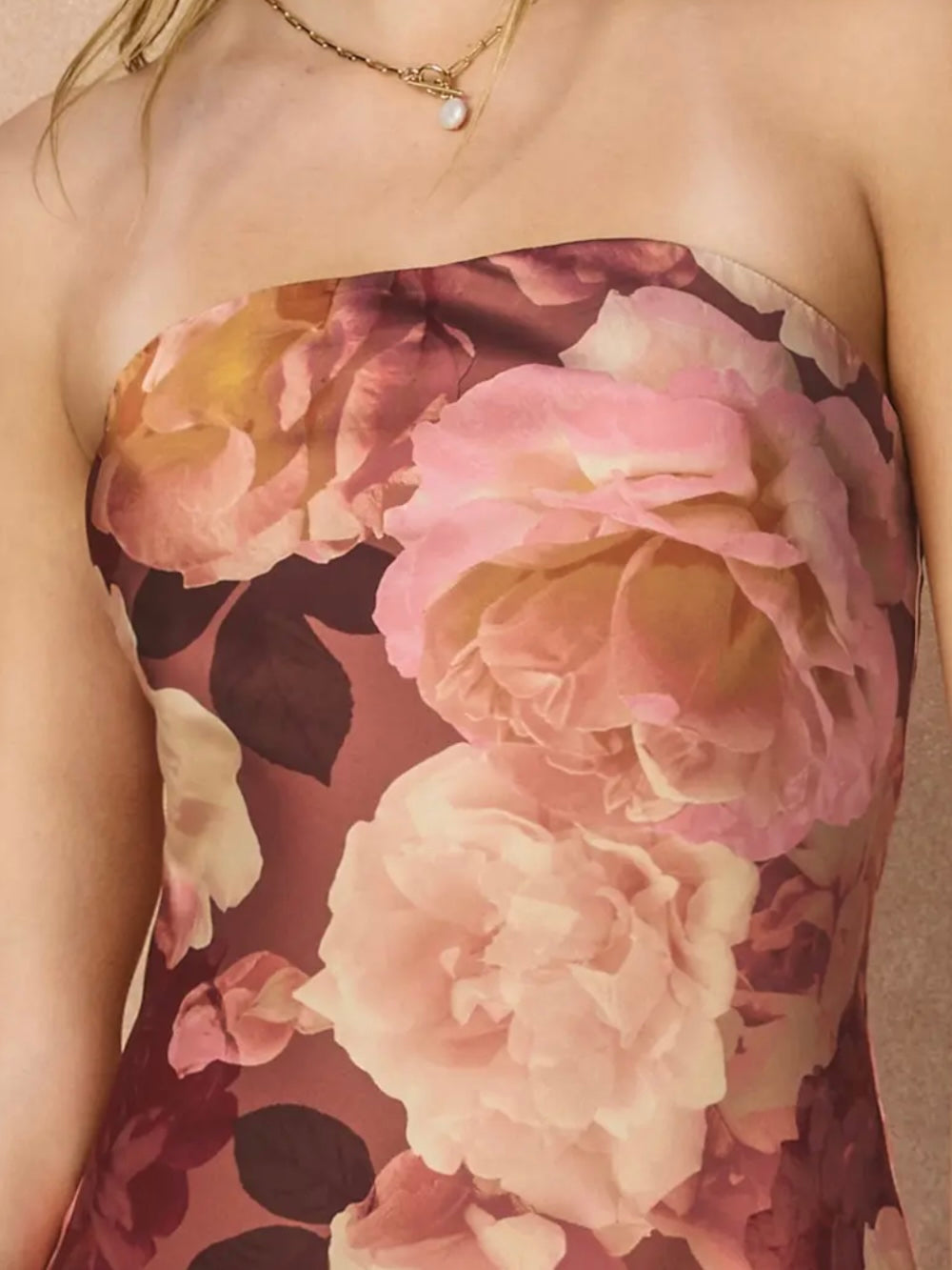 Close-up of a strapless floral top or dress with large pink and cream rose print on a mauve-brown background, worn by a model with a delicate gold necklace.