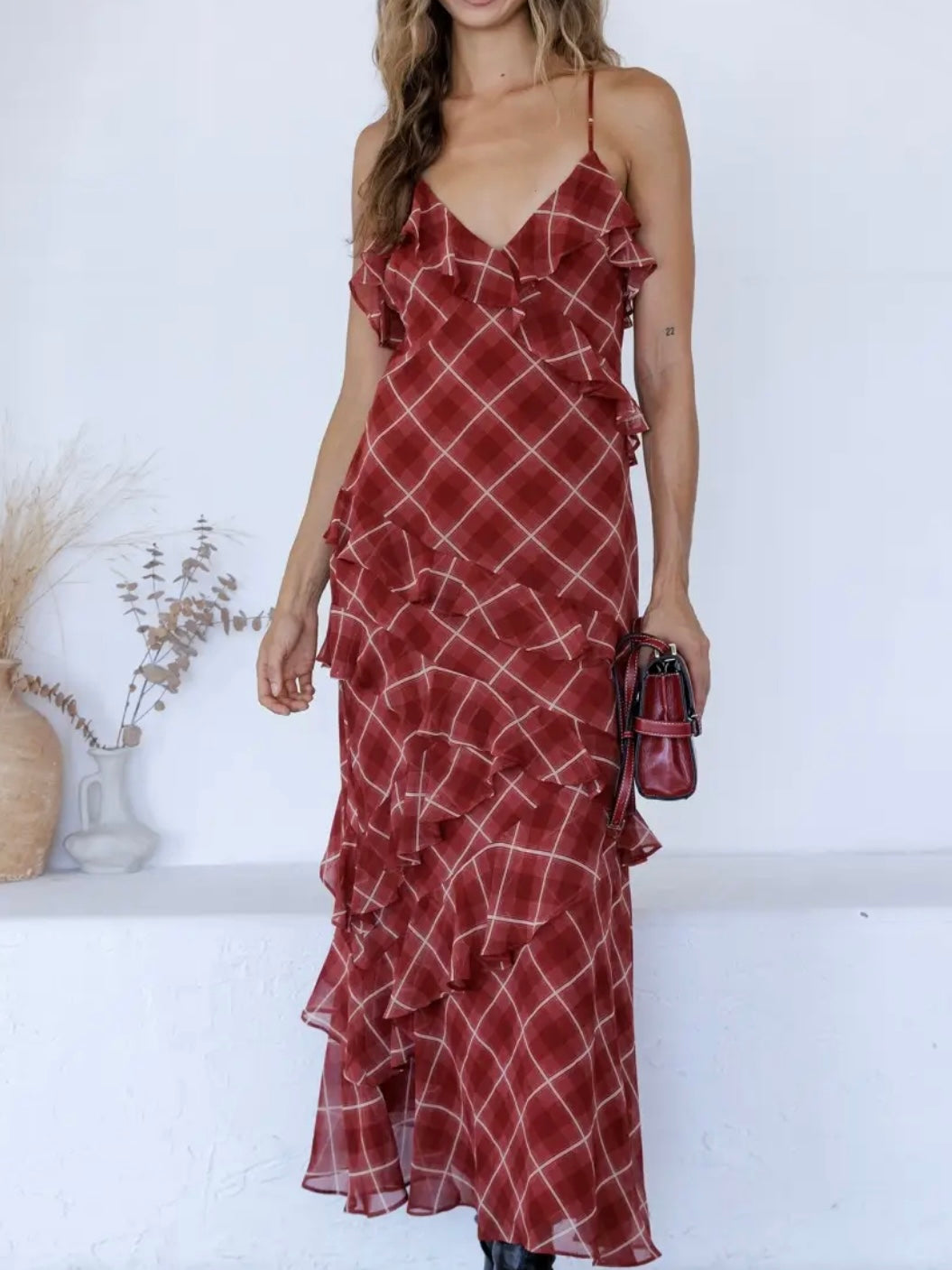 A woman wearing a red ruffled spaghetti-strap maxi dress with a white grid pattern, holding a matching red handbag against a white wall background.
