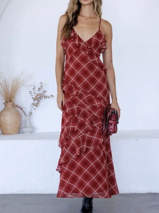 A woman models a red and white plaid maxi dress with ruffle details, holding a matching red handbag in a bright, minimalist room with white walls and decorative vases.