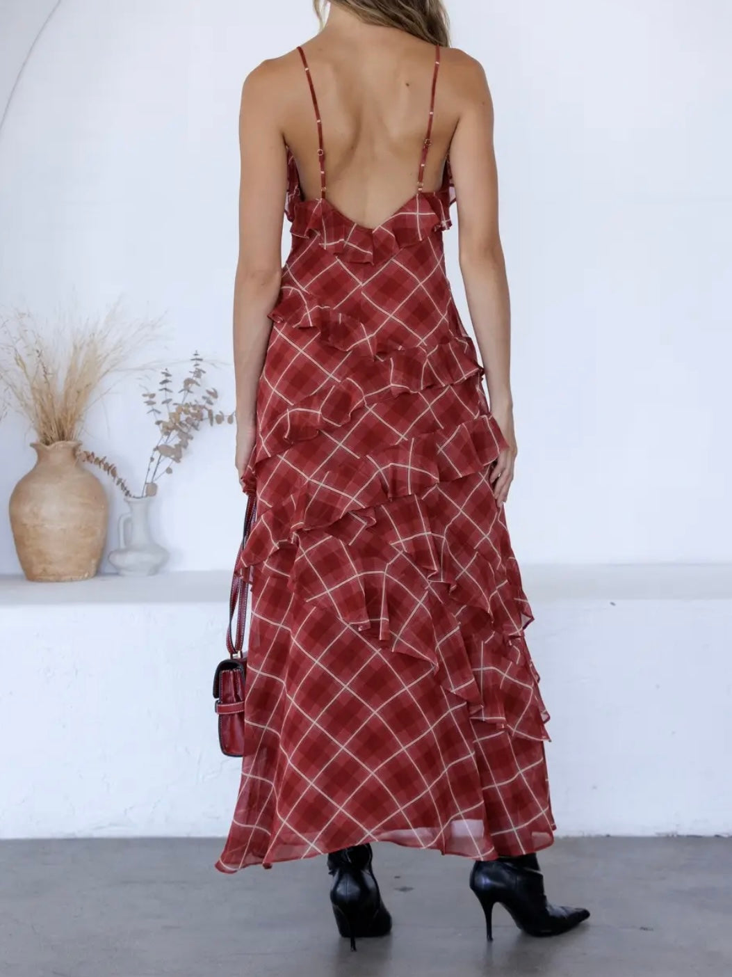 A woman shown from the back wearing a red and white plaid maxi dress with thin straps and a ruffled tiered skirt, paired with black high-heeled boots and a small red handbag, in a bright white indoor setting.