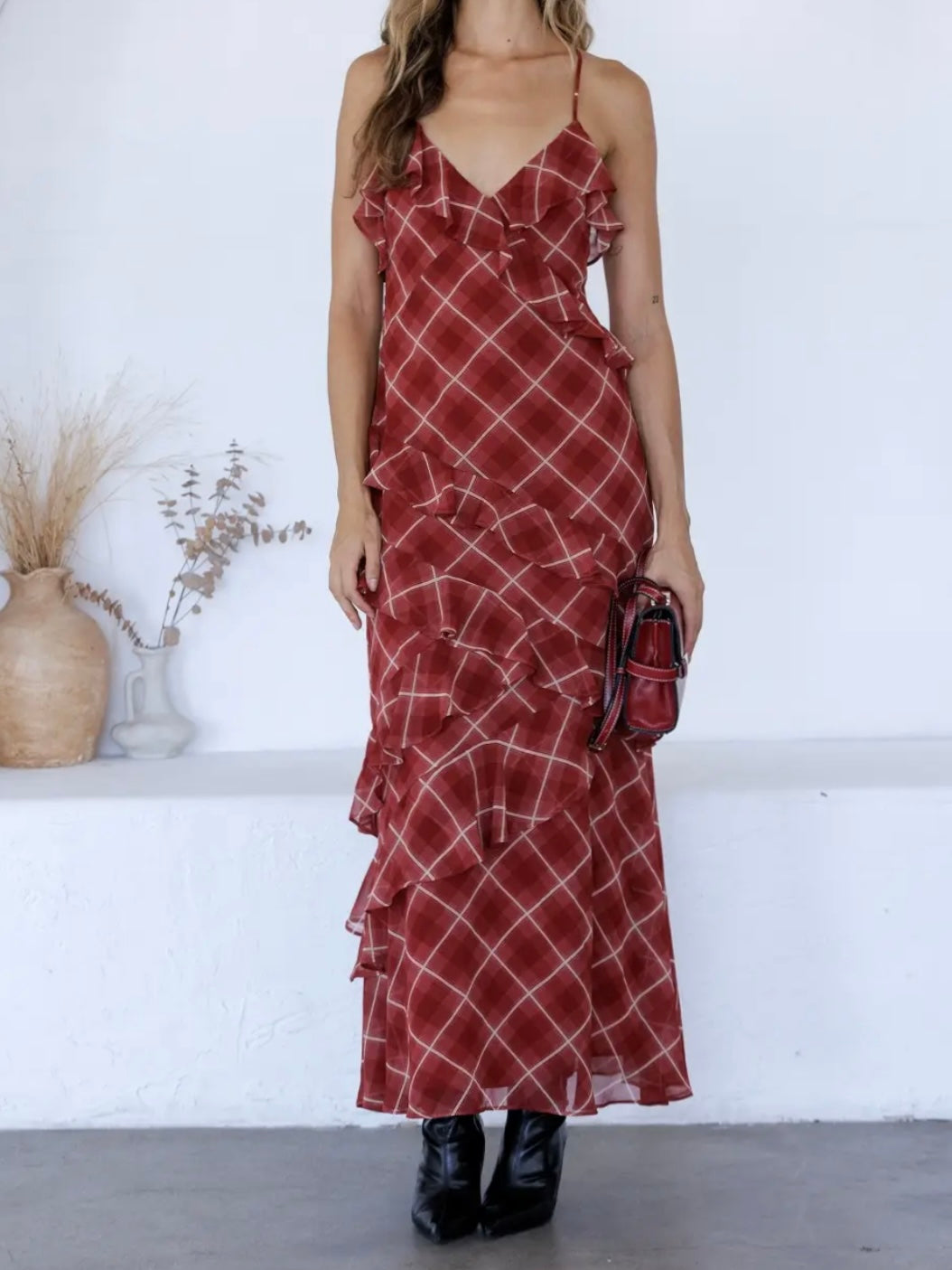 A woman models a rust-red maxi dress with a white plaid pattern and ruffled details, paired with black ankle boots and a matching small handbag, set against a white wall with decorative dried plants.
